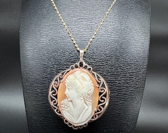 May include: A gold-toned necklace featuring a cameo pendant. The pendant showcases a carved profile of a woman in white against an orange background, framed by an ornate, openwork border. The necklace is displayed on a black mannequin.