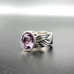 May include: A silver ring with a woven band design. The ring features a large, oval-shaped purple gemstone. The gemstone is set in a bezel setting, and the band has a textured, braided appearance.