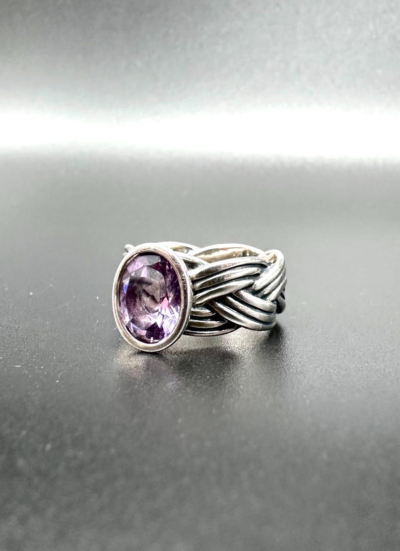 May include: A silver ring with a woven band design. The ring features a large, oval-shaped purple gemstone. The gemstone is set in a bezel setting, and the band has a textured, braided appearance.
