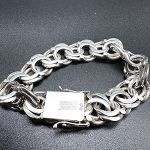 Puede incluir: Una pulsera de plata con un diseño de cadena gruesa. La pulsera presenta eslabones ovalados entrelazados y un cierre rectangular. El cierre está grabado con el texto "NIEMA L 830S Y9". La pulsera se muestra sobre una superficie oscura.