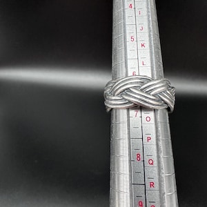 May include: A silver-colored ring with a woven design is displayed on a ring sizer. The ring is made of a shiny metal and has a textured surface. The ring is approximately a size 7.