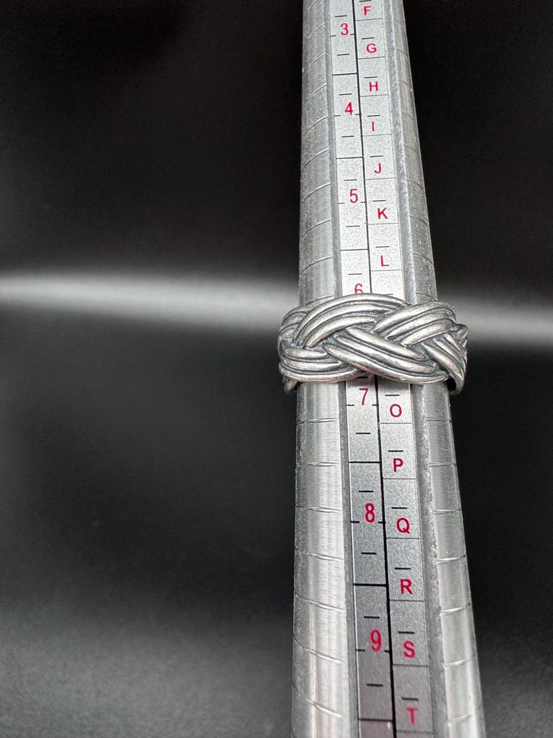 May include: A silver-colored ring with a woven design is displayed on a ring sizer. The ring is made of a shiny metal and has a textured surface. The ring is approximately a size 7.