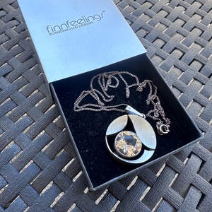 May include: A silver-toned pendant necklace with a large, faceted, amber-colored crystal in a circular setting. The necklace is displayed in a black box with the text "finnfeelings SCANDINAVIAN DESIGN" on the lid.