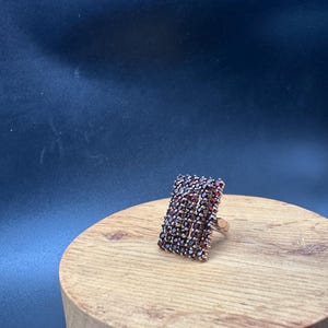 Könnte beinhalten: Ein rechteckiger Ring mit einer dichten Ansammlung kleiner, tiefroter Granat-Edelsteine, die in ein goldfarbenes Metallband gefasst sind. Der Ring hat eine markante, mehrreihige Anordnung von Steinen, die die gesamte Oberseite bedeckt.