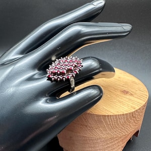 Puede incluir: Un anillo tipo cluster con numerosas piedras pequeñas de color rojo intenso y talladas, engastadas estrechamente en un marco metálico. La cabeza ovalada del anillo está densamente cubierta por estas gemas rojas, creando una superficie texturizada.
