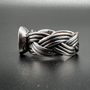 May include: A silver ring with a woven band design. The ring features a large, oval-shaped stone set in a bezel. The band is intricately braided, creating a textured appearance. The ring is displayed against a dark background.