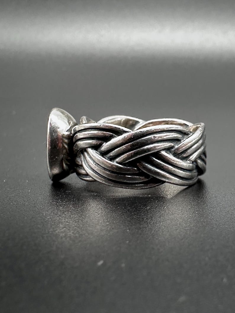 May include: A silver ring with a woven band design. The ring features a large, oval-shaped stone set in a bezel. The band is intricately braided, creating a textured appearance. The ring is displayed against a dark background.