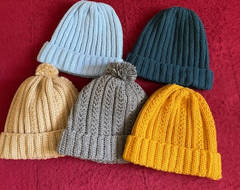 May include: Five knitted beanies in various colors: light blue, teal, mustard yellow, tan, and gray. The beanies have a ribbed knit design, with one tan beanie and one gray beanie featuring a pom-pom.