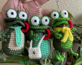 May include: Three handmade crochet frog keychains. Each frog is green with large white eyes, a red tongue, and a silver keychain. One frog has a green tie, another a white purse, and the third a yellow scarf.