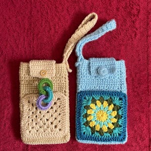 May include: Two handmade crochet phone pouches. One is beige with a button closure and a multi-colored ring accent. The other is light blue with a floral square design and a button closure. Both have a strap.