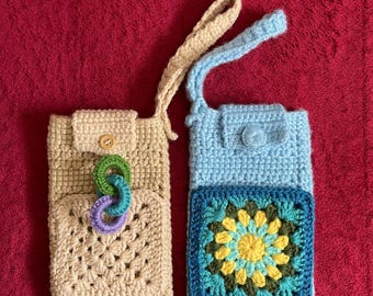 May include: Two handmade crochet phone pouches. One is beige with a button closure and a multi-colored ring accent. The other is light blue with a floral square design and a button closure. Both have a strap.