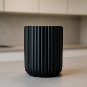 Könnte beinhalten: Eine schwarze, zylindrische Vase mit vertikalen, geriffelten Details. Die Vase hat einen abgerundeten Boden und eine leicht ausgestellte Oberseite. Die matte Oberfläche und das schlichte Design verleihen ihr eine moderne Ästhetik. Die Vase eignet sich für die Inneneinrichtung.