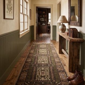May include: A long rectangular area rug featuring a geometric pattern in shades of olive green, brown, and cream. The rug has a fringed edge and is positioned on a wooden floor in a hallway with green wainscoting, a wooden console table, and boots.