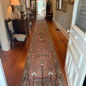 May include: A long, narrow runner rug featuring a dusty rose background with a repeating symmetrical pattern in shades of sage green, cream, and dark blue. The rug spans the length of a wooden hallway floor, extending from the foreground to the doorway.