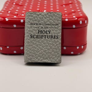 Pode incluir: Um pequeno livro cinza, intitulado "New World Translation of the Holy Scriptures", está em frente a uma lata vermelha com bolinhas brancas. O livro tem uma capa texturizada e está centralizado.