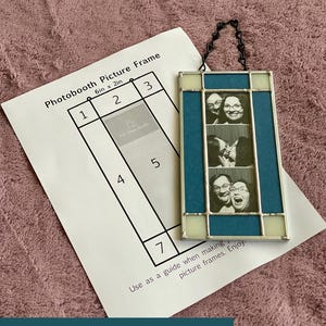Puede incluir: Un marco de fotos rectangular de vidrieras con paneles de vidrio azul y crema, que cuenta con una cadena de metal negro para colgar. El marco contiene una tira vertical de tres fotos en blanco y negro. Se incluye una guía de papel etiquetada como Photobooth Picture Frame 6in x 2in.