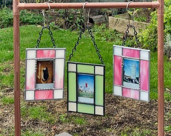 May include: Three stained glass photo frames hanging from a copper pipe structure. The frames are in pink, green, and white, each with a different photo inside. The frames are suspended by black chains, set against a green lawn.