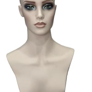 Pode incluir: Busto de manequim com maquilhagem. O manequim tem um tom de pele neutro, com maquilhagem detalhada nos olhos em tons de preto, prata e verde. Os lábios são rosa suave. Uma touca de peruca bege é visível na cabeça.