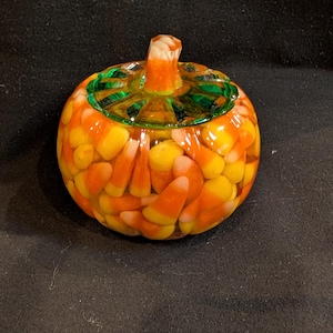 May include: A round, pumpkin-shaped resin sculpture filled with numerous candy corn pieces. The top features a green, translucent, ribbed lid with a small, orange and white candy corn-shaped stem protruding from the center.