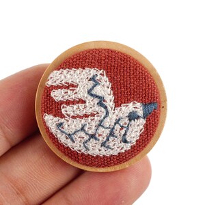May include: A round, wooden pin with a red fabric center. The fabric is embroidered with a white and blue bird design. The pin is held in a hand.