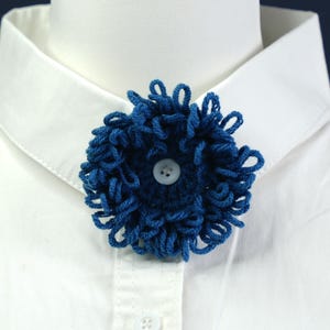 Może przedstawiać: Ręcznie robiona, ciemnoniebieska broszka w kształcie kwiatu z szydełka z białym guzikiem pośrodku, przypięta do białej koszuli z kołnierzykiem. Broszka wykonana jest z pętelkowej przędzy, tworząc trójwymiarowy, teksturowany wzór kwiatowy. Kołnierz koszuli jest czysty.