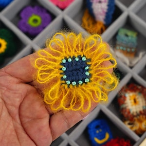 May include: A handmade yellow and blue crochet sunflower with a dark blue center and light blue bead accents. The sunflower is held in a hand, with other crochet flowers in the background.