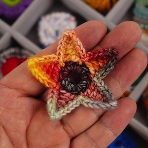 May include: A handmade, five-pointed star-shaped crochet item. The star features a gradient of colors, including yellow, orange, red, and brown, with a dark center. The crochet stitch is visible, and the item is held in a hand.