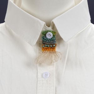 May include: A colorful, handmade brooch with a button closure, attached to a white collared shirt. The brooch features green, blue, and orange yarn, with small beads and a fringe detail. A second button is visible on the shirt.