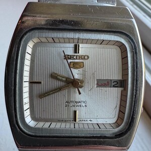 May include: A vintage Seiko 5 automatic watch with a silver rectangular face and a stainless steel strap. The watch has a silver dial with gold hands and markers, a day-date display, and the text "SEIKO 5 AUTOMATIC 21 JEWELS JAPAN".