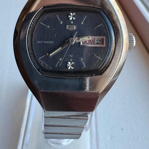 May include: A vintage Seiko automatic watch with a silver case and a dark blue dial. The watch face shows the day and date, with silver hands and markers. The watch is displayed on a clear stand.