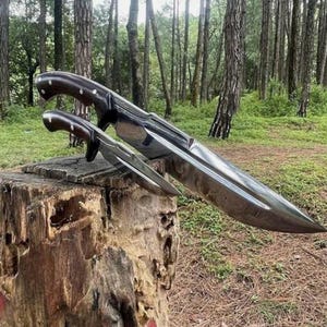 May include: Two knives with polished steel blades and dark wooden handles rest on a tree stump. The larger knife has a long, curved blade, while the smaller one has a shorter, more pointed blade. The handles are dark brown with silver rivets.