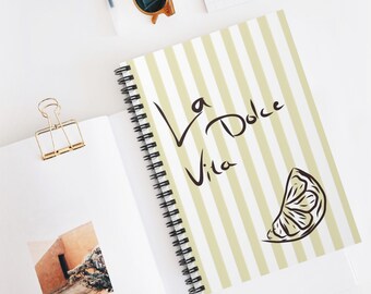 Op de afbeelding: Een spiraalgebonden notitieboek met een kaft met verticale strepen in lichtgeel en wit. De kaft bevat de tekst "La Dolce Vita" in zwart schrift, samen met een citroenpartje illustratie. Andere items zijn zonnebrillen en een gouden paperclip.