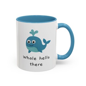 Op de afbeelding: Witte keramische mok met een blauw interieur en handvat. De mok heeft een cartoon walvis illustratie in blauwtinten, met de tekst "Whale hello there" eronder.