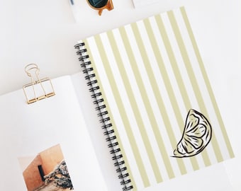 Op de afbeelding: Een spiraalgebonden notitieboek met een kaft met verticale strepen in lichtgeel en wit. Een zwart-wit illustratie van een citroenpartje staat op de kaft. Andere items op een wit oppervlak zijn een gouden paperclip en een zonnebril.