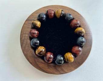 May include: A bracelet made of polished tiger eye beads in shades of red, gold, and blue. The beads are arranged in a circular pattern, resting on a black velvet surface within a wooden bowl.