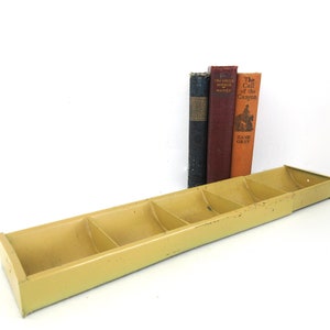 May include: A yellow metal shelf with six compartments, designed to hold books or other items. The shelf is empty, but there are three books stacked behind it. The books are titled "The Creek Mirror", "The Call of the Canyon", and "Zane Grey".
