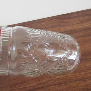 May include: A clear glass jar with a red lid, featuring a decorative embossed pattern. The jar is lying on a wooden surface, showcasing its detailed design and color contrast.