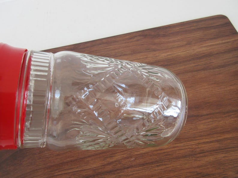 May include: A clear glass jar with a red lid, featuring a decorative embossed pattern. The jar is lying on a wooden surface, showcasing its detailed design and color contrast.