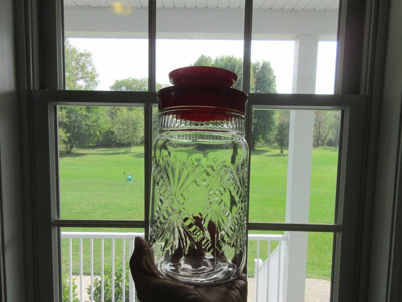 May include: A clear glass jar with a red lid, held in front of a window. The jar has a decorative, diamond-shaped pattern. The background shows a green lawn and trees, suggesting a vintage or antique kitchen storage item.