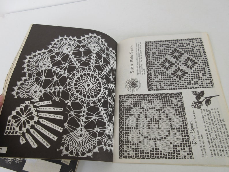 K&ouml;nnte beinhalten: Ein Vintage-H&auml;kelbuch mit Anleitungen zum H&auml;keln eines Spinnennetz-Quadrats, eines Rosenquadrats und einer Doily. Das Buch hat schwarze Seiten und wei&szlig;e H&auml;kelmuster.
