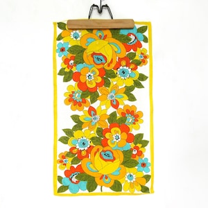 May include: A rectangular fabric towel featuring a vibrant floral pattern with large yellow, orange, and turquoise flowers accented by dark green leaves on a white background, finished with a solid yellow border and held by a wooden hanger.