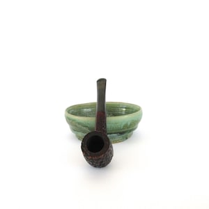 May include: A dark brown tobacco pipe with a textured bowl and a dark stem, resting in front of a small, light green ceramic bowl. The pipe has a classic shape and design, suitable for tobacco use.