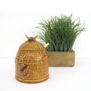 May include: A ceramic honey pot shaped like a beehive with a bee on the lid. The pot is brown and yellow with a textured surface. It comes with a small wooden honey dipper.