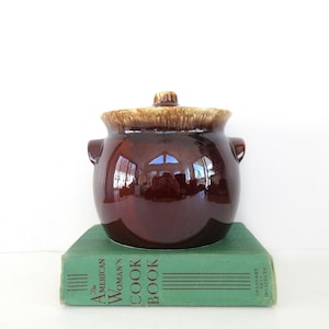 May include: A vintage brown ceramic bean pot with a lid, sitting on a green cookbook. The pot has a glossy finish and two side handles. The lid is a lighter brown color. The cookbook reads "The American Woman's Cook Book".