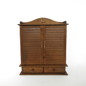 May include: A vintage wooden cabinet with a dark brown finish. The cabinet features two louvered doors, two small drawers, and a decorative carved element at the top. The cabinet has a classic, antique aesthetic.