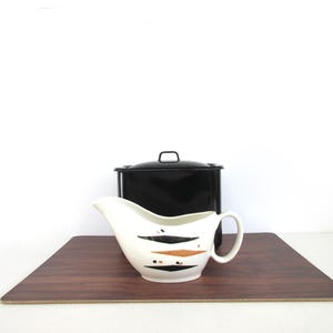 May include: A white ceramic gravy boat with a black, brown, and orange abstract design. A black rectangular casserole dish with a lid is behind the gravy boat. The items are on a brown wooden surface.