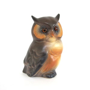 May include: A ceramic owl figurine with brown and orange painted details. The owl has large, round, orange eyes.