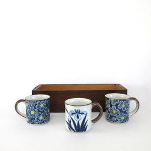 May include: Three ceramic mugs with brown handles and floral designs. Two mugs feature a blue floral pattern with yellow centers, while the central mug displays a blue iris design. A wooden box is behind the mugs.
