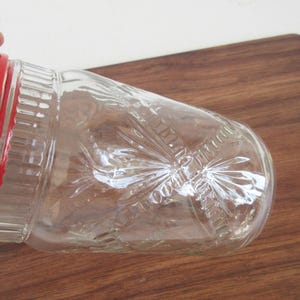 May include: Clear glass jar with a red lid. The jar has a decorative pattern and is partially visible. The jar is on a wooden surface.