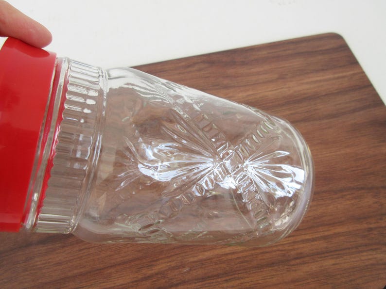 May include: Clear glass jar with a red lid. The jar has a decorative pattern and is partially visible. The jar is on a wooden surface.